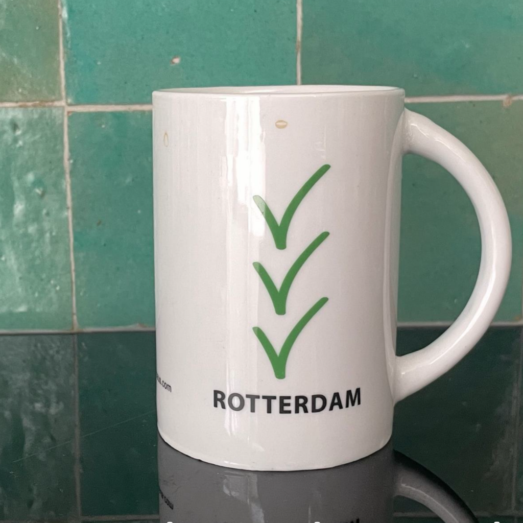 Een witte beker, aan twee kanten bedrukt, de ene kant met 'Amsterdam' en de andere 'Rotterdam'. Dit is de "Rotterdam"-kant, met 3 groene vinkjes. Want in Rotterdam kan het wel, zoals iedereen weet.